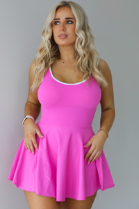 Woman wearing a hot pink athleisure dress that has white trim, a fitted bodice, and flowy skirt. The photo shows a closeup of the dress. The photo is taken indoors against a white wall.
