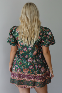 A woman shown from behind wearing a dark green dress that has a mauve and multicolored floral print pattern throughout, has puffed sleeves, a collared neckline with functional gold button closures in the front, standing indoors against a white wall.