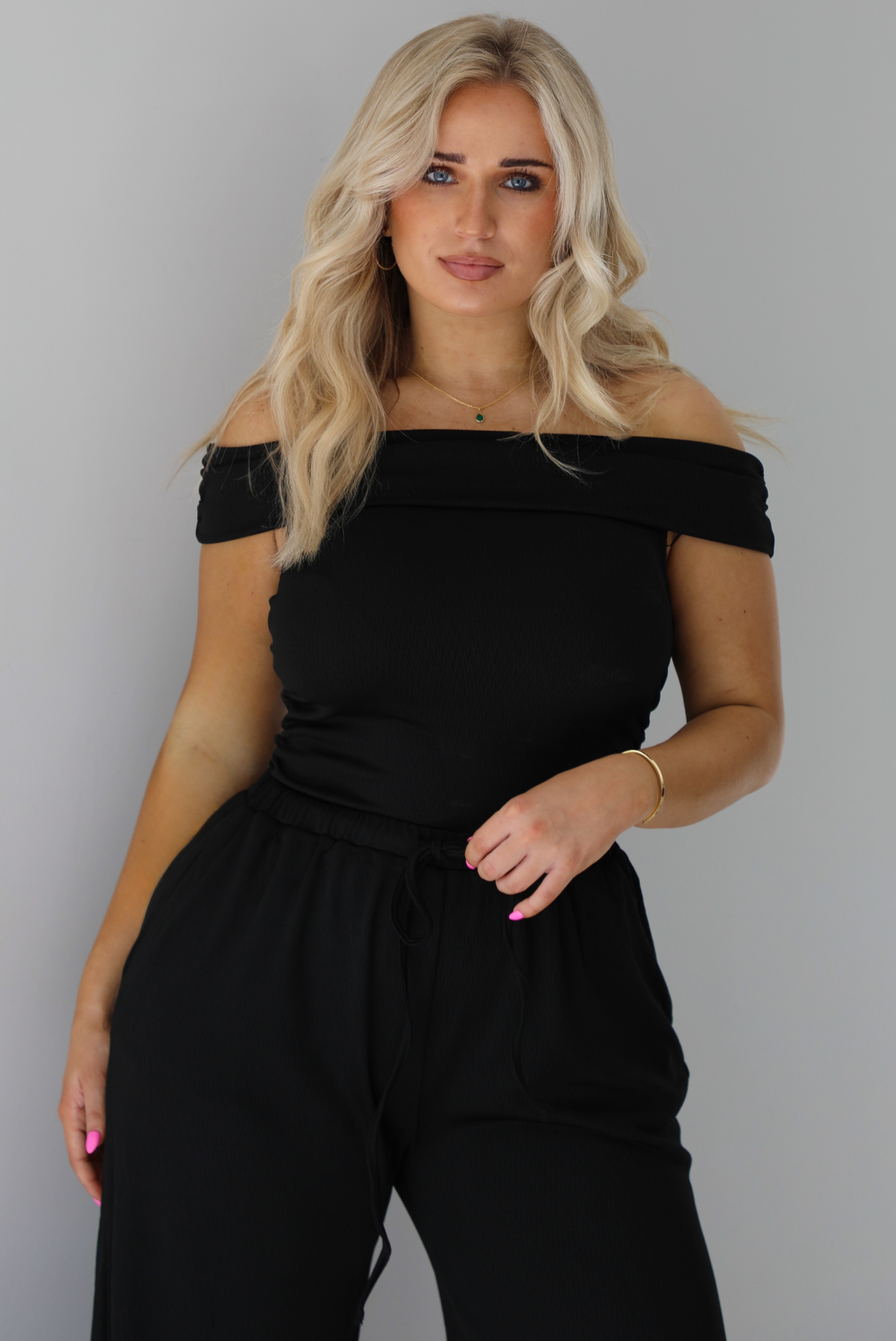 Woman wearing a two piece set featuring an off the shoulder top and high waisted pants. The photo shows a closeup of the top. The photo is taken indoors against a white wall.