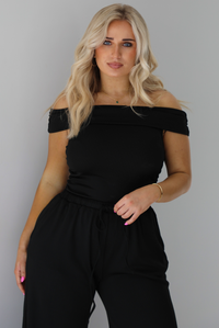 Woman wearing a two piece set featuring an off the shoulder top and high waisted pants. The photo shows a closeup of the top. The photo is taken indoors against a white wall.