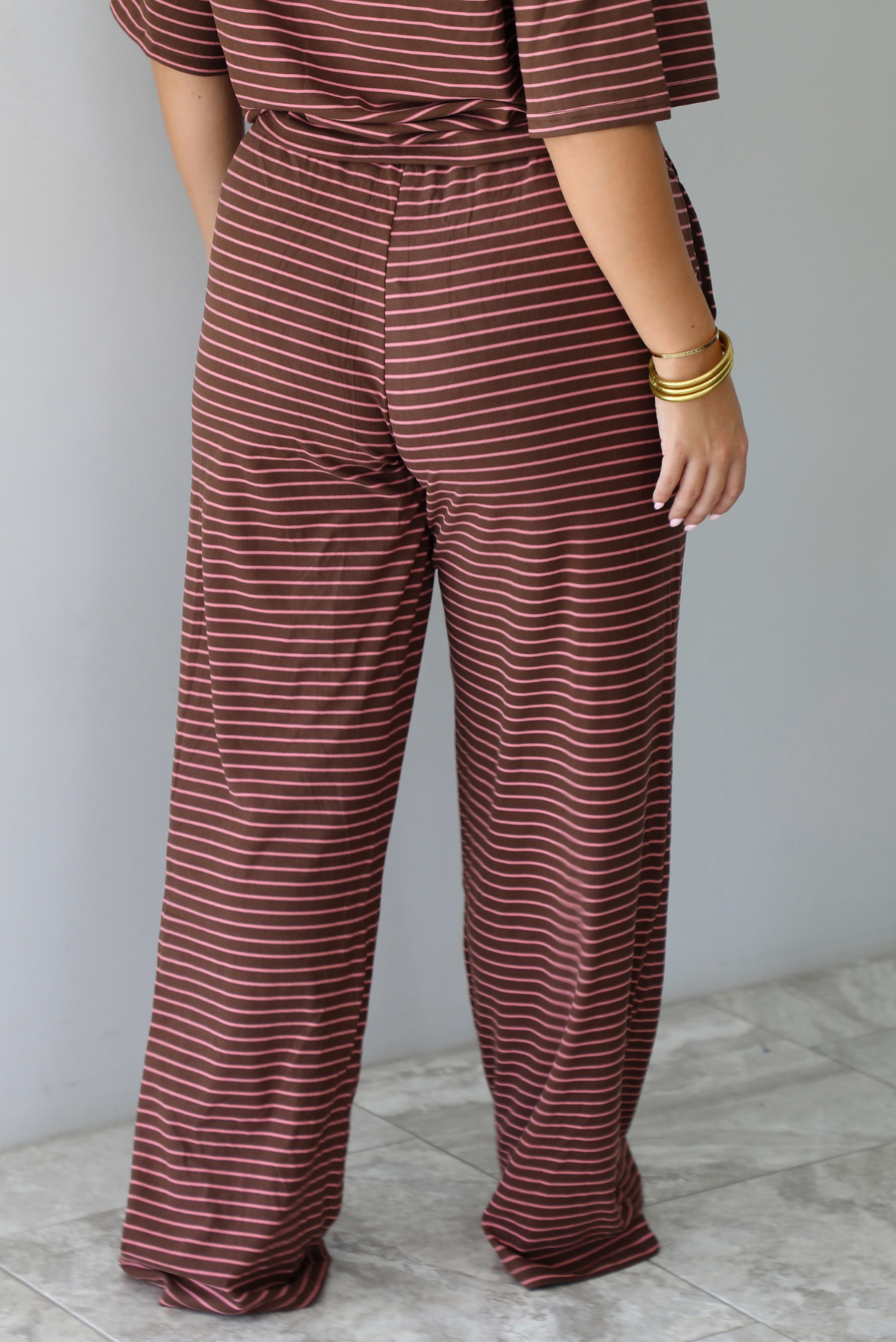 A woman shown from behind wearing a brown and pink striped two piece set featuring a slightly cropped tee and high waisted pants, standing indoors against a white wall with white marble flooring. The photo shows a closeup of the back of the pants, which have an elastic waistline and functional pockets on either side. 