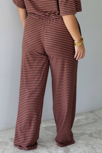 A woman shown from behind wearing a brown and pink striped two piece set featuring a slightly cropped tee and high waisted pants, standing indoors against a white wall with white marble flooring. The photo shows a closeup of the back of the pants, which have an elastic waistline and functional pockets on either side. 