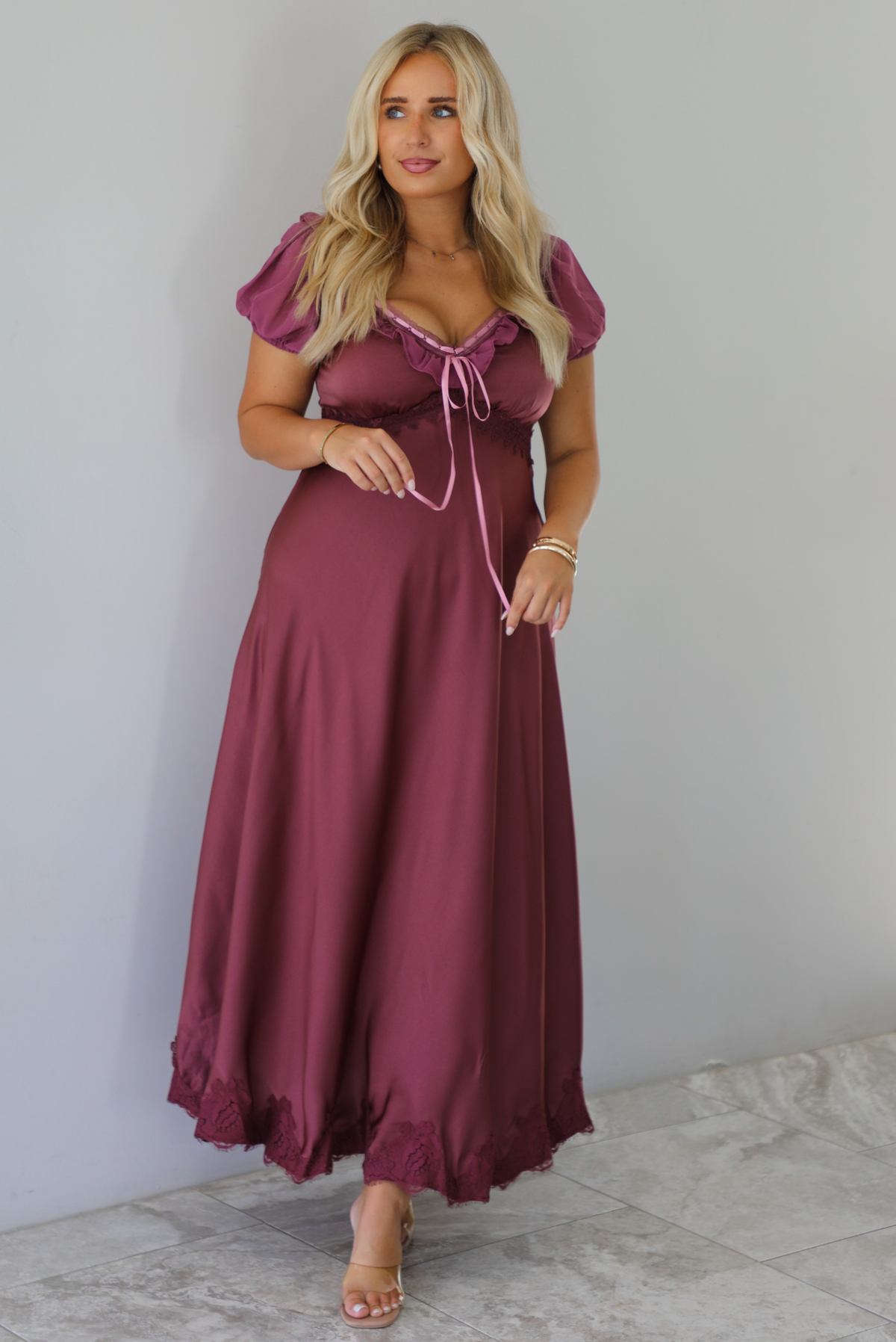A woman modeling a long, burgundy maxi dress with short puff sleeves, a lace-up sweetheart neckline, and a flowing satin-like skirt, standing indoors against a white wall with white marble flooring. 