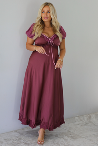 A woman modeling a long, burgundy maxi dress with short puff sleeves, a lace-up sweetheart neckline, and a flowing satin-like skirt, standing indoors against a white wall with white marble flooring. 