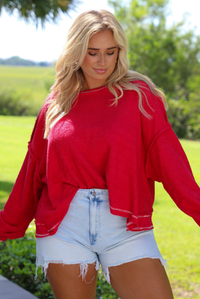 Woman wearing an oversized long sleeved sweater with light wash denim shorts outdoors with greenery in the background. 