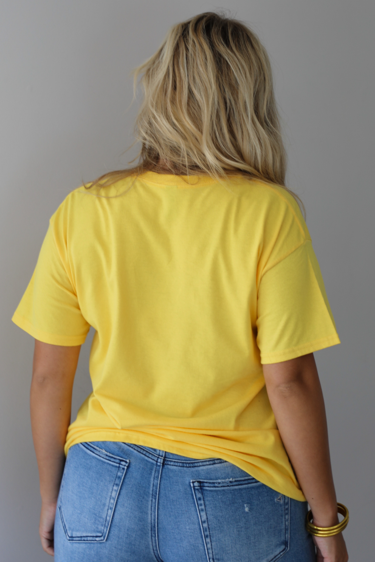 A woman shown from behind wearing a bright yellow graphic t-shirt with blue sardine illustrations and the text 'Sardines LISBON FISH MARKET', paired with blue jeans, standing indoors against a white wall.
