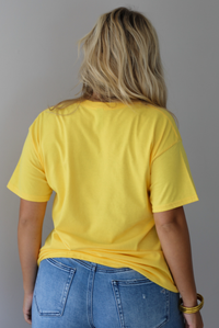 A woman shown from behind wearing a bright yellow graphic t-shirt with blue sardine illustrations and the text 'Sardines LISBON FISH MARKET', paired with blue jeans, standing indoors against a white wall.