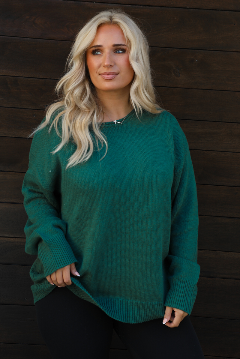Woman model wearing a green oversized sweater with black leggings, white socks, and tan Uggs. The photo is taken outdoors against a dark brown door. The photo is taken up close and showcases the sweater. 