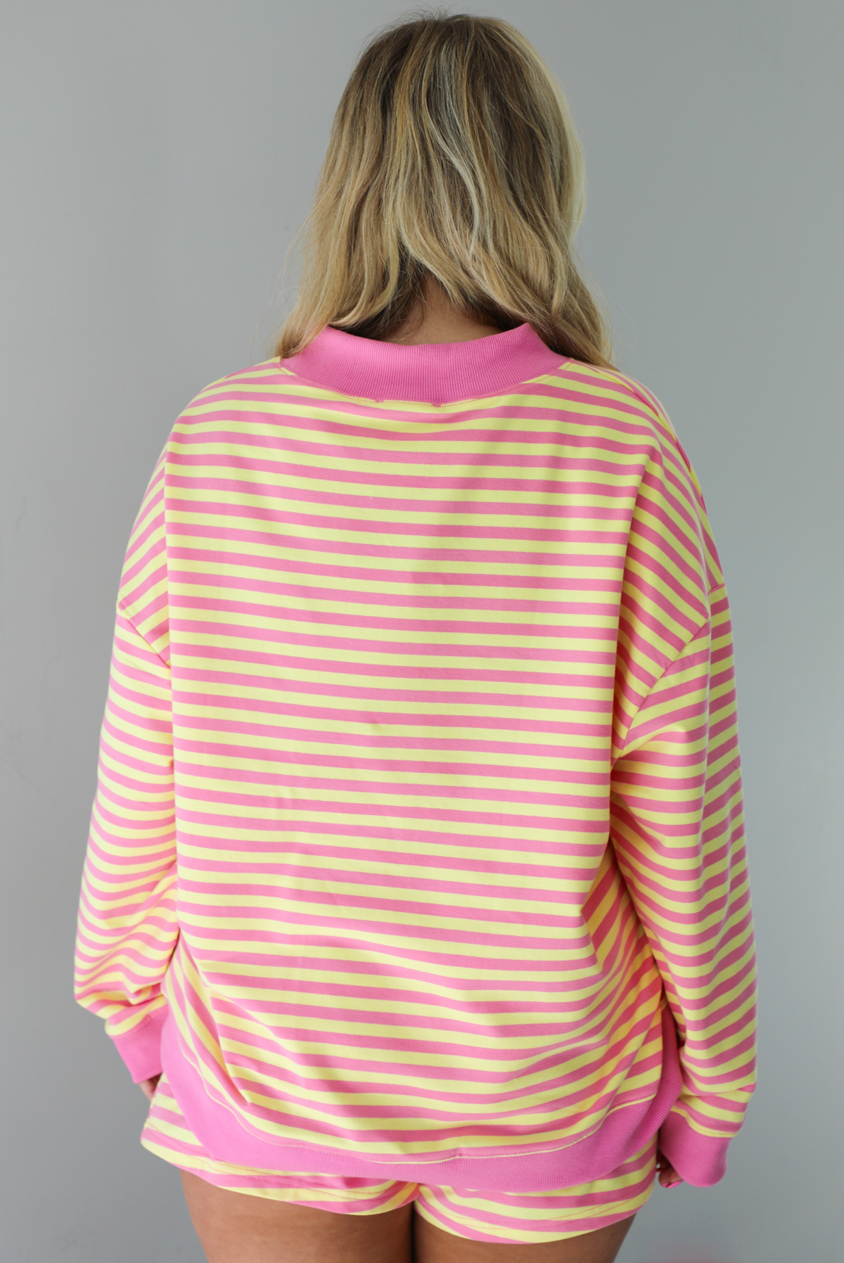 Woman wearing a pink and yellow striped set featuring an oversized sweatshirt and shorts. The photo is taken indoors against a white wall and shows a closeup of the back of the top.