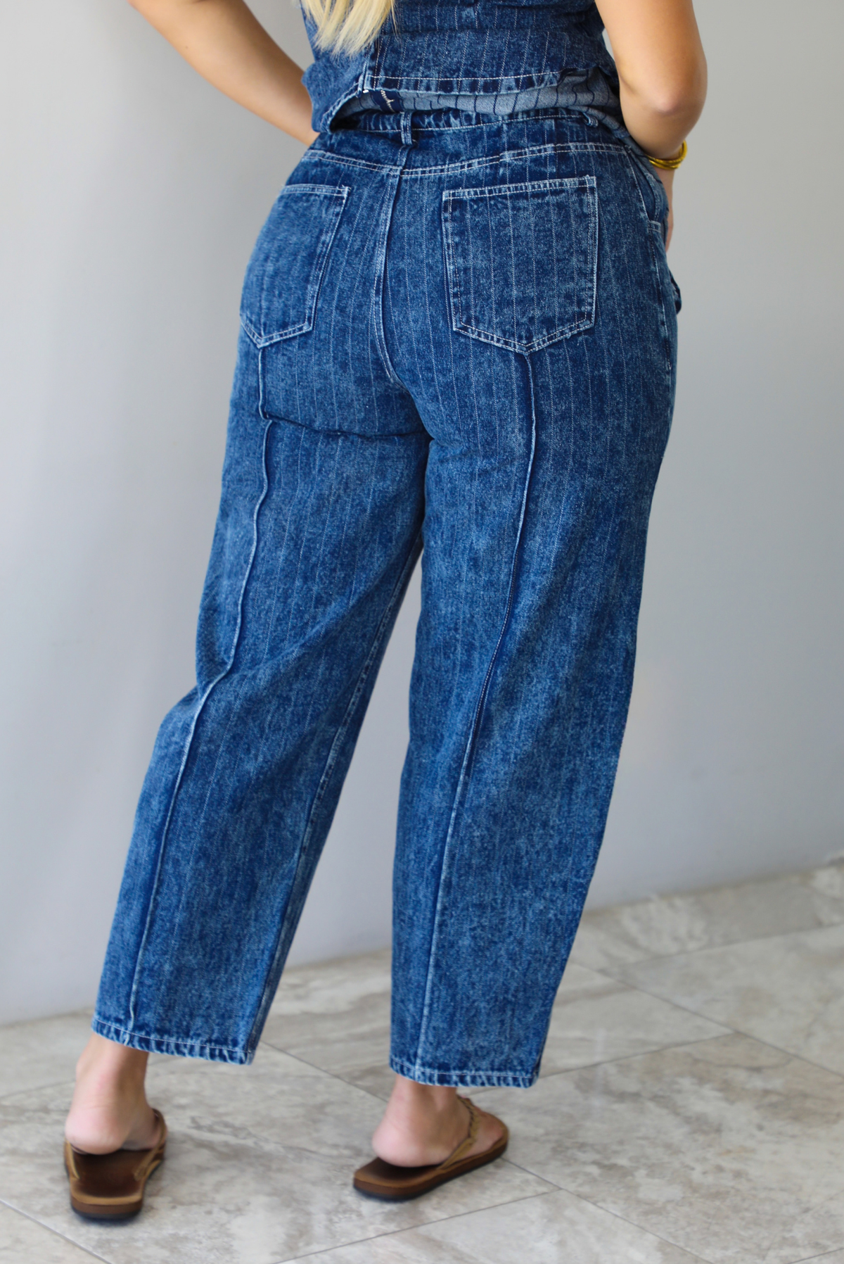 A woman shown from behind wearing a matching two piece dark wash denim set with a pin striped design on it featuring a sleeveless peplum style top and high waisted denim jeans, standing indoors against a white wall with white marble flooring. The photo focuses on the back of the pants only, which have functional back pockets. 