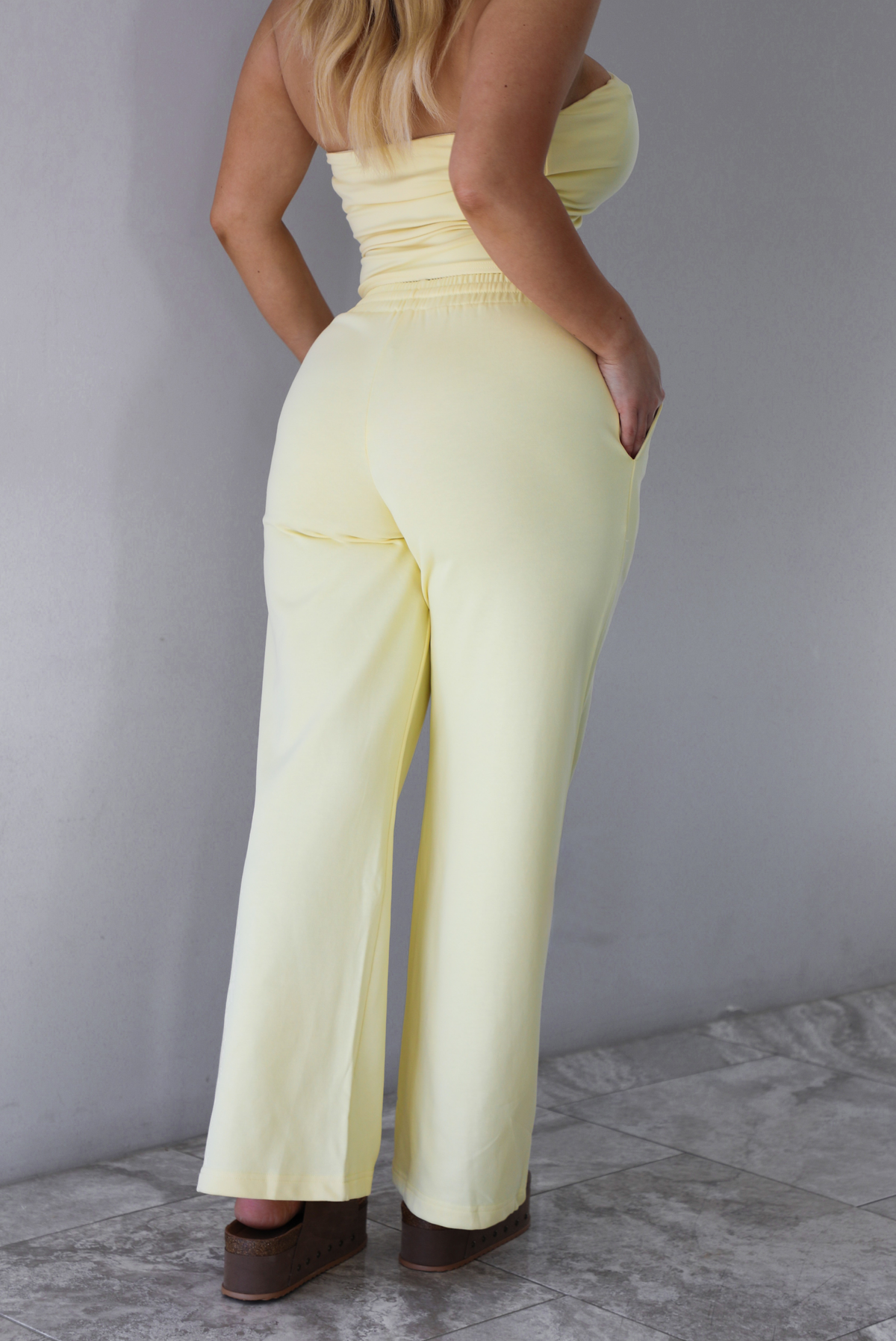 Woman wearing a pale yellow two piece set featuring a strapless tube top and high waisted, flared hem pants that have an elastic and drawstring waistline with functional pockets on either side. The photo is taken indoors against a white wall with white marble flooring and shows a closeup of the back of the pants.