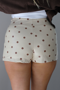 A woman shown from behind wearing a cream mini skort with a brown polka dot print pattern throughout, standing indoors against a white wall.