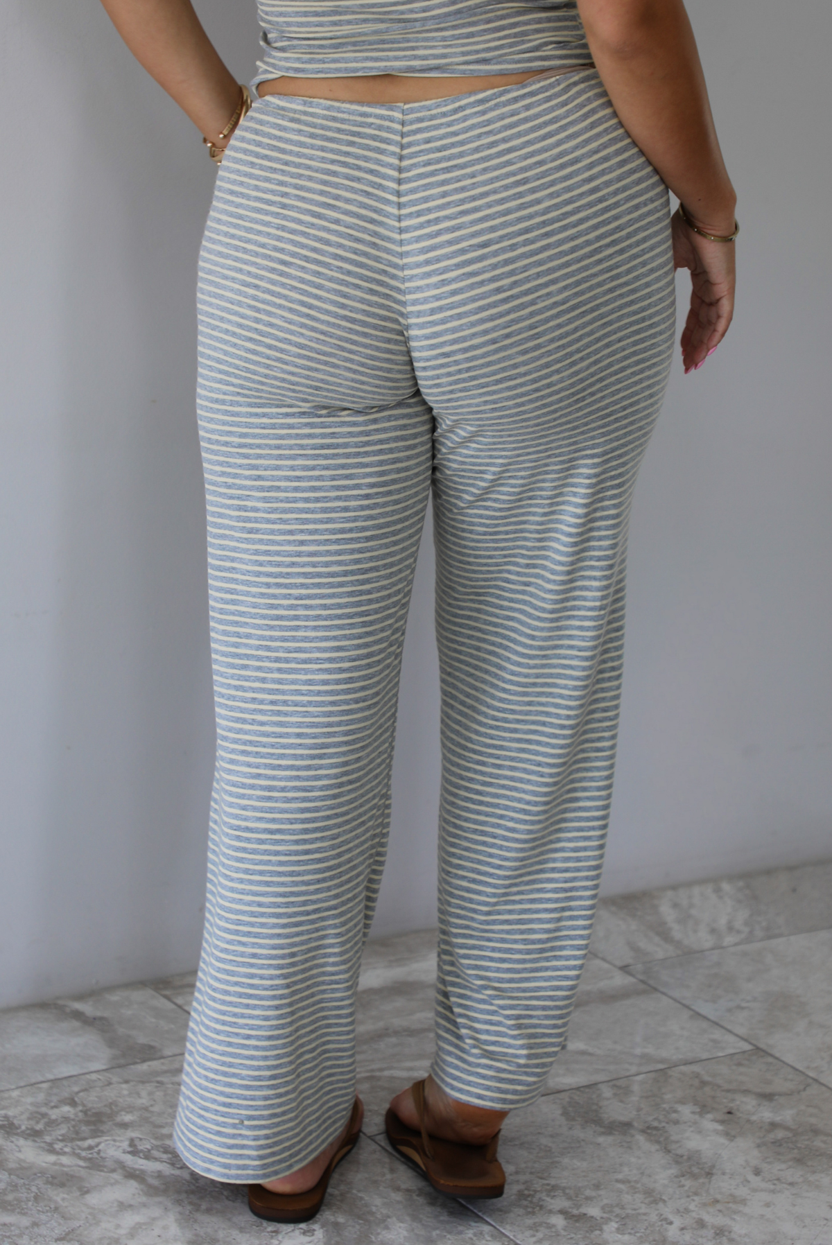 A person shown from behind wearing light gray and pale yellow horizontally striped wide-leg pants with a drawstring waistband, paired with brown sandals, standing indoors against a white wall with white marble flooring.