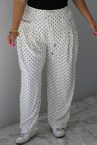 A closeup of a woman shown from the waist down wearing sand white pants with a black polka dot print pattern throughout, they have a drawstring waistline, functional pockets on either side, and pleated details at the hips to give it a barrel effect, standing indoors against a white wall with white marble flooring. 