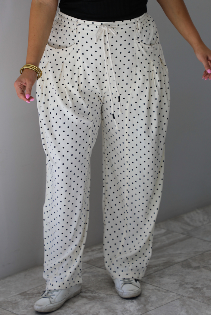 A closeup of a woman shown from the waist down wearing sand white pants with a black polka dot print pattern throughout, they have a drawstring waistline, functional pockets on either side, and pleated details at the hips to give it a barrel effect, standing indoors against a white wall with white marble flooring. 