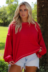 Woman wearing an oversized long sleeved sweater with light wash denim shorts outdoors with greenery in the background. 