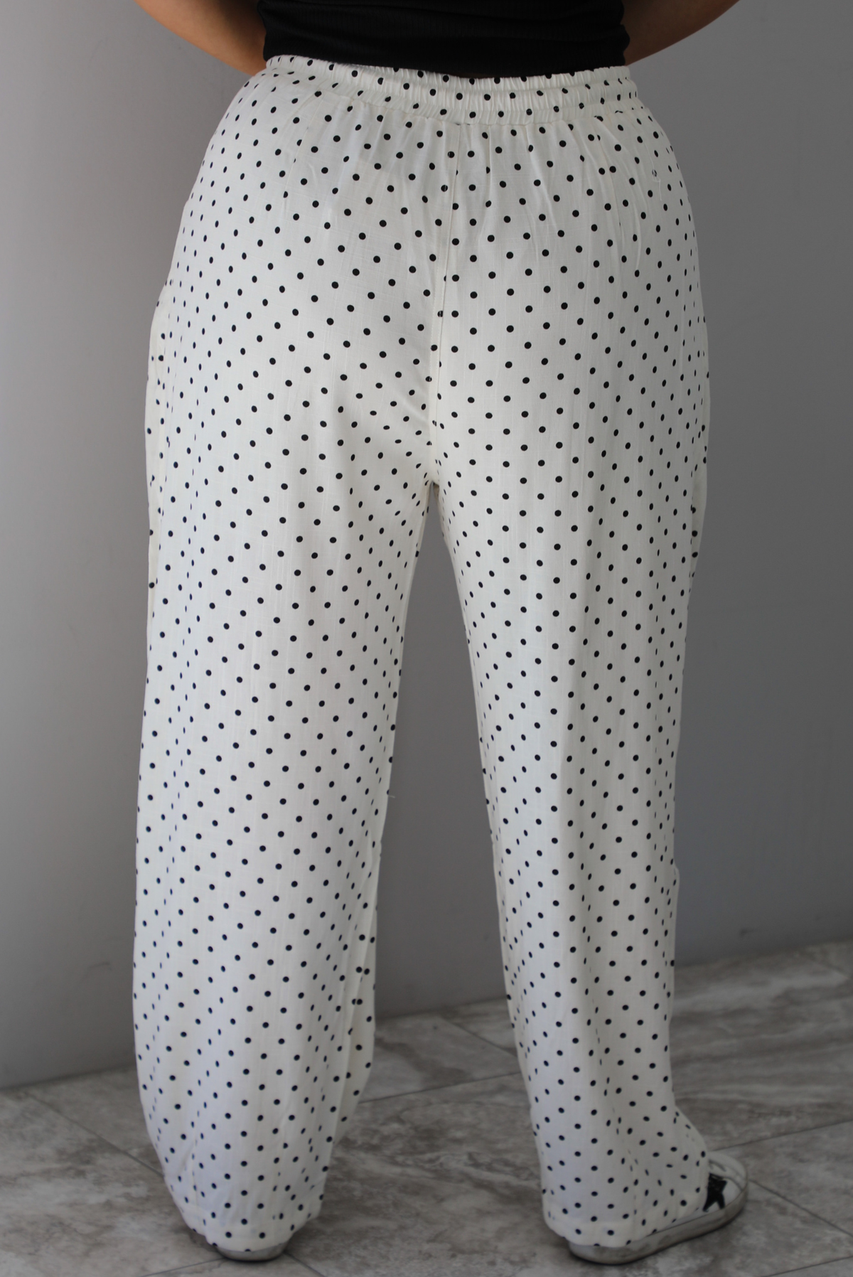 A closeup of a woman shown from behind from the waist down wearing sand white pants with a black polka dot print pattern throughout, they have a drawstring waistline, functional pockets on either side, and pleated details at the hips to give it a barrel effect, standing indoors against a white wall with white marble flooring. 