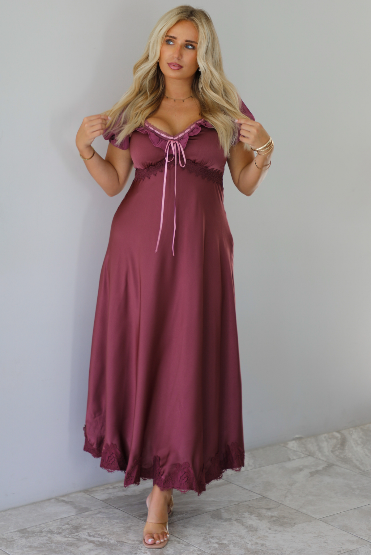 A woman modeling a long, burgundy maxi dress with short puff sleeves, a lace-up sweetheart neckline, and a flowing satin-like skirt, standing indoors against a white wall with white marble flooring. 