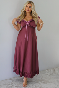 A woman modeling a long, burgundy maxi dress with short puff sleeves, a lace-up sweetheart neckline, and a flowing satin-like skirt, standing indoors against a white wall with white marble flooring. 