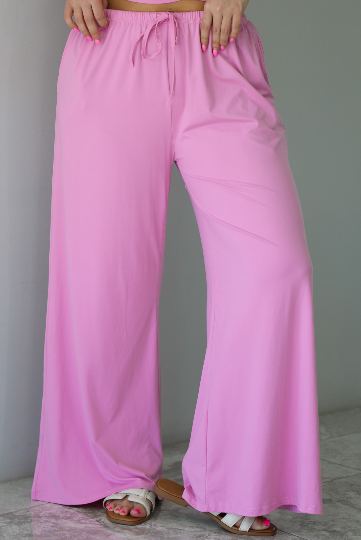 Woman wearing a two pieced pink set featuring a halter top and comfy pants with a flared hem detail. The photo is taken indoors against a white wall with white marble flooring, and shows a closeup of the pants, which have an elastic and drawstring waistline with functional pockets on either side. 