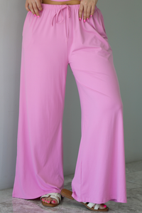 Woman wearing a two pieced pink set featuring a halter top and comfy pants with a flared hem detail. The photo is taken indoors against a white wall with white marble flooring, and shows a closeup of the pants, which have an elastic and drawstring waistline with functional pockets on either side. 