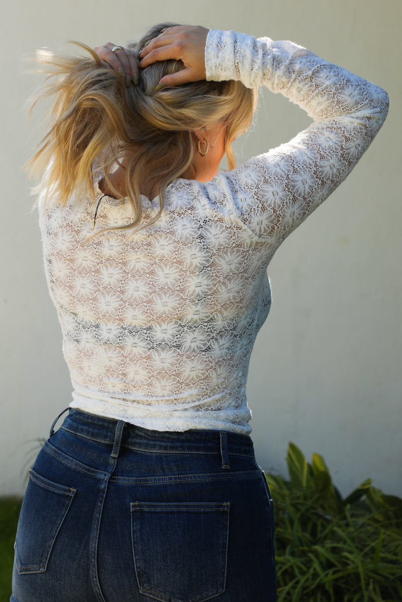 Close up of the back of a model wearing white sheer lace top with a floral pattern with dark denim jeans outside.