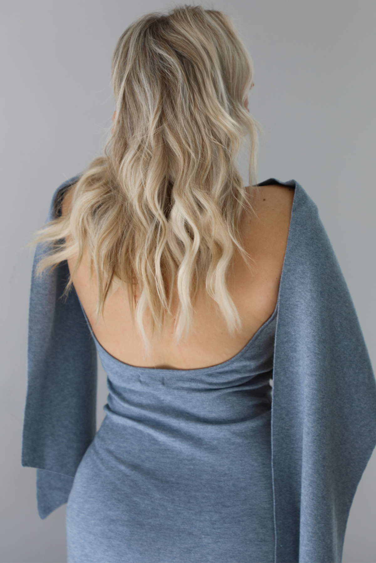 Woman wearing a grey maxi length knit dress that has a high neckline with an attached shawl detail that covers the upper half of the chest and flows on either shoulder in the back. The photo shows the back of the dress, which has a low scoop back cut out. The photo is taken indoors against a white wall.