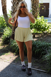 A woman modeling a white tank and pale yellow shorts that have a brown polka dot pattern throughout, standing outdoors with palm trees and other greenery in the background. 