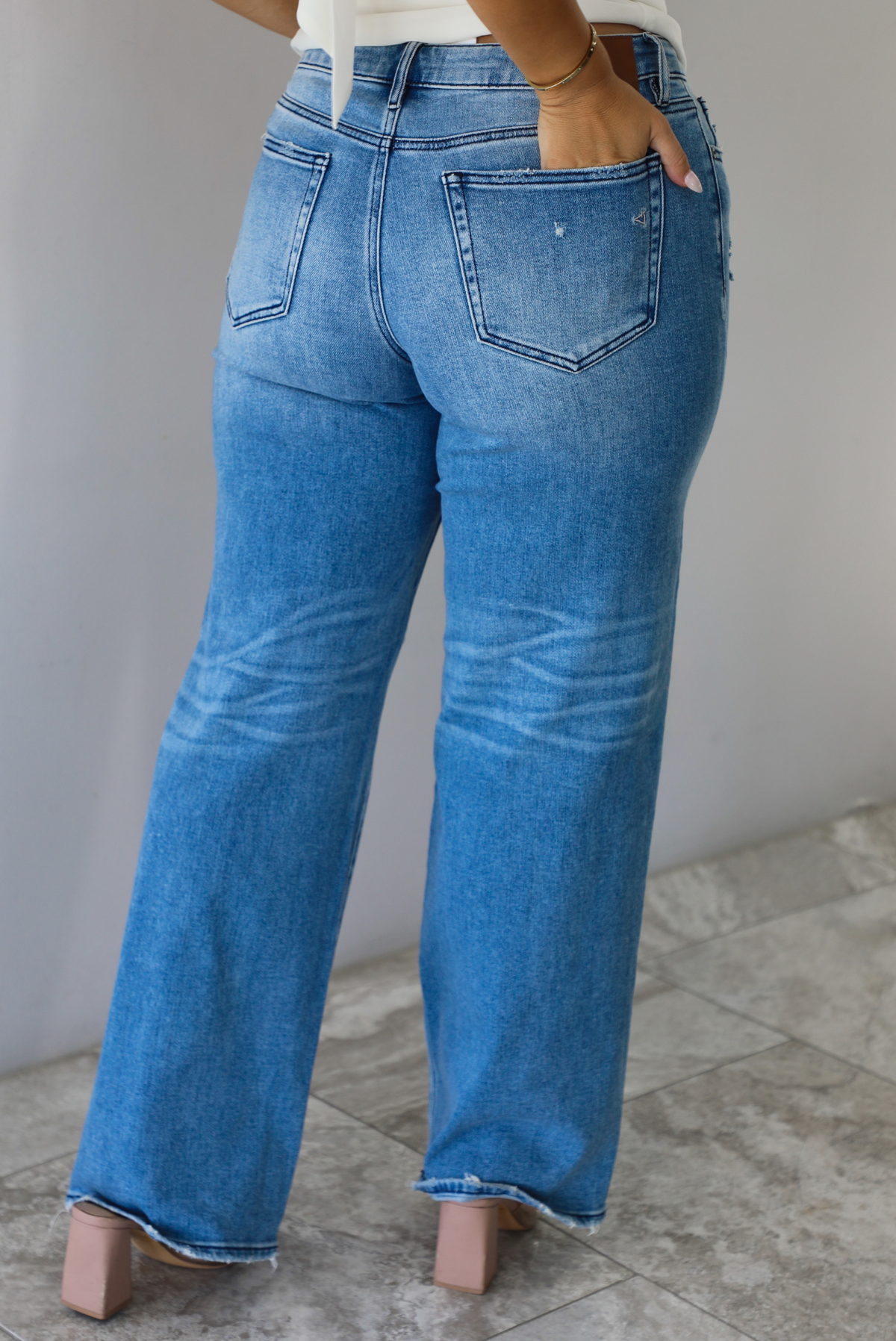 Closeup of a woman from behind wearing mid-wash denim jeans with a mid-rise waistline, distressed details, and flared hems indoors against a white wall with white marble flooring. 