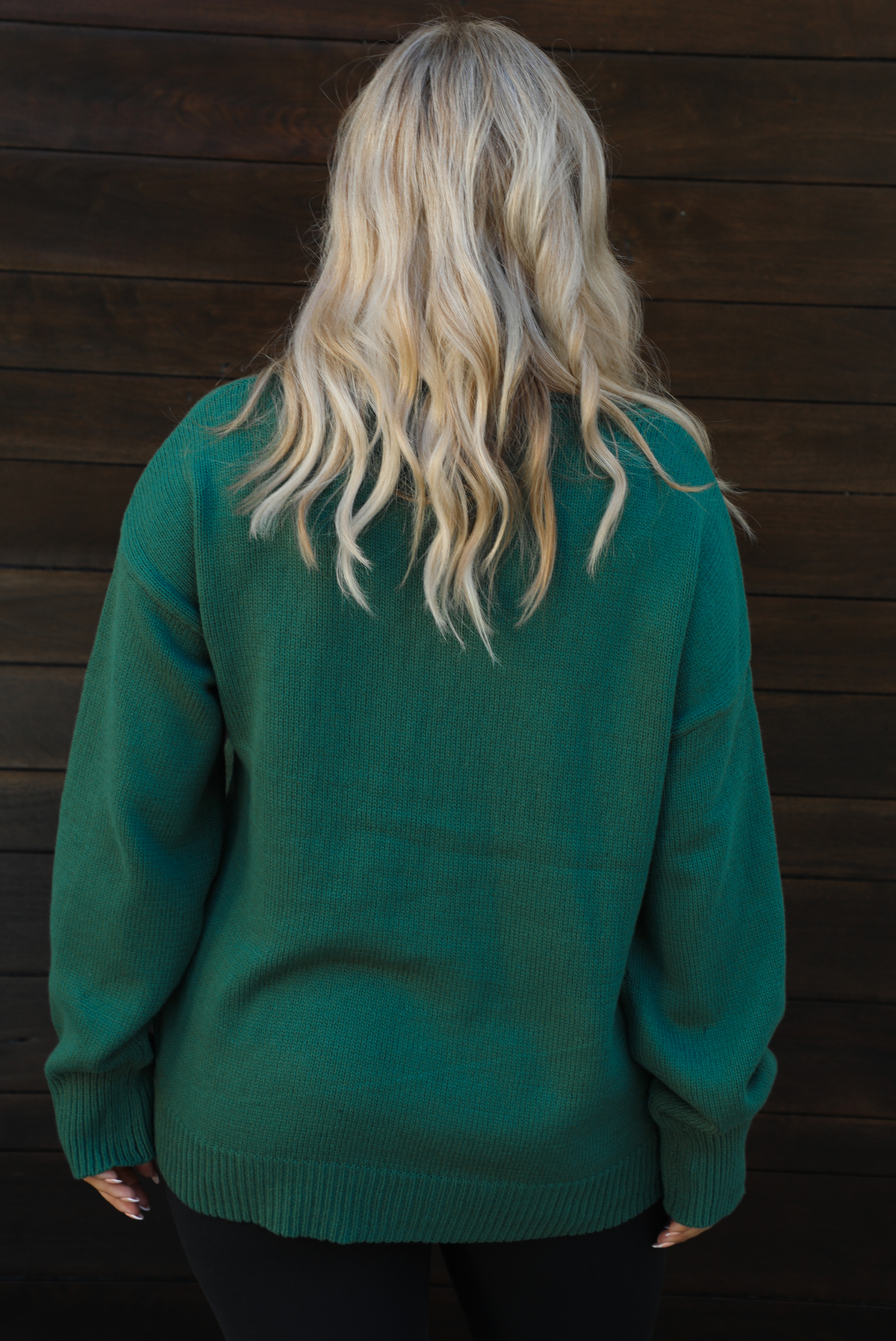 Woman model wearing a green oversized sweater with black leggings, white socks, and tan Uggs. The photo is taken outdoors against a dark brown door. The photo is taken up close and showcases the back of the sweater. 