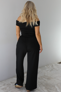 Woman wearing a two piece set featuring an off the shoulder top and high waisted pants. The photo shows the back of the set.  The photo is taken indoors against a white wall with white marble flooring.