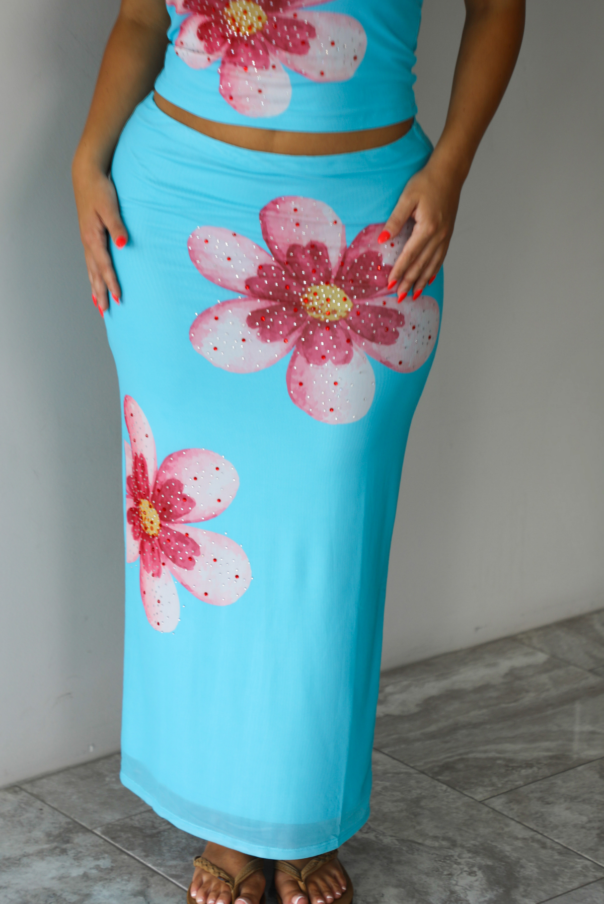 A blonde woman modeling a bright turquoise blue set with large pink and white hibiscus flower prints, indoors against a white wall with white marble flooring. The photo shows a closeup of the skirt, which is high waisted with a maxi length. 