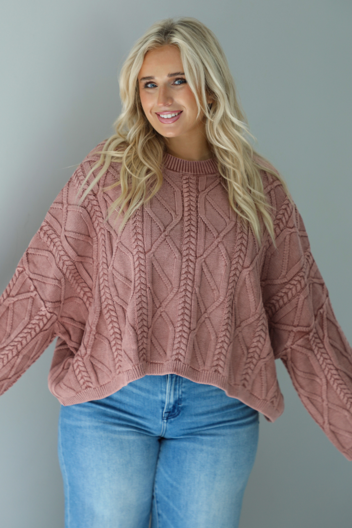A woman wearing a mulberry oversized sweater paired with a pair of medium wash distressed denim, standing indoors against a white wall.