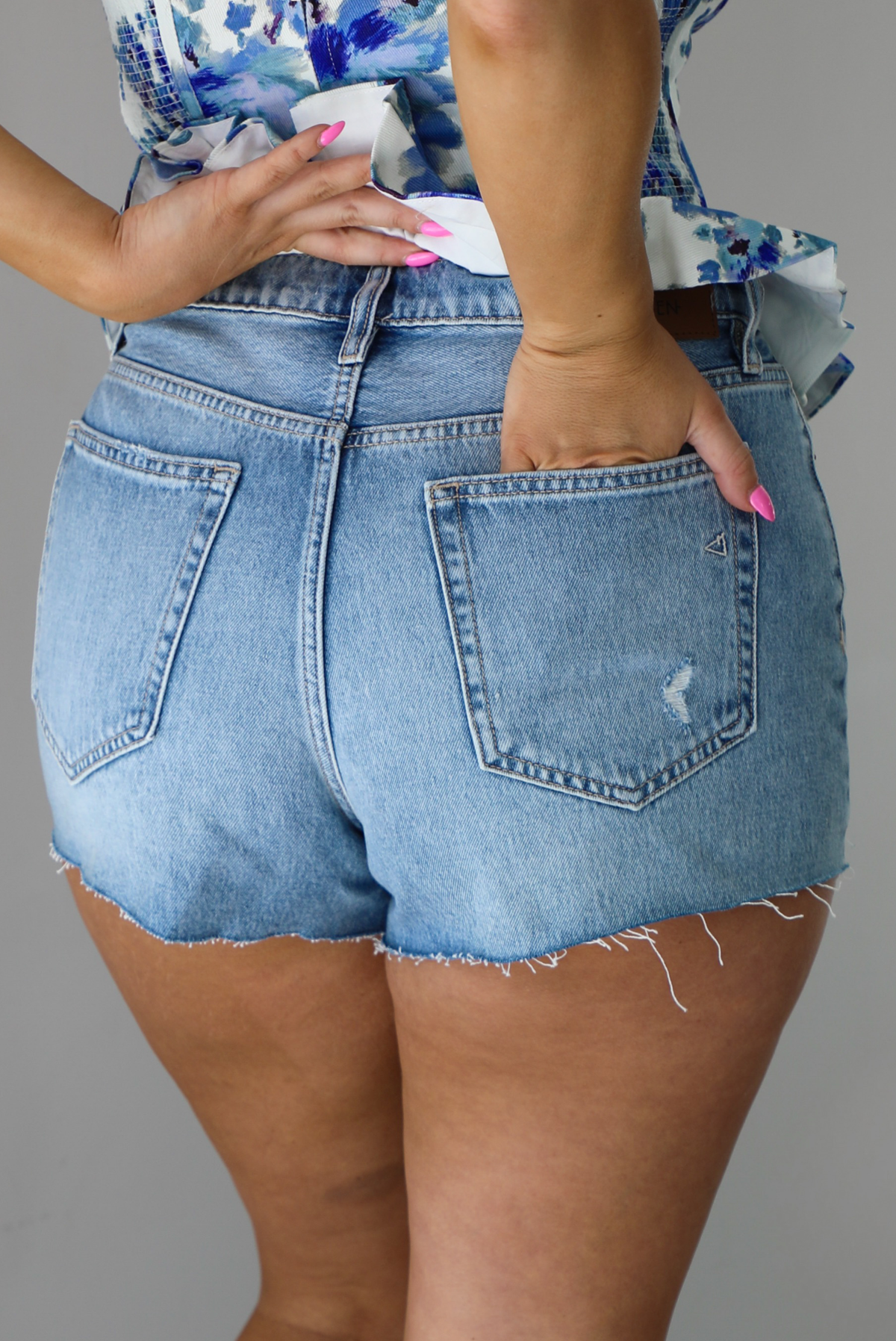 Woman wearing a light-wash pair of denim shorts that have distressed detailing. The photo shows the back of the shorts. The photo is taken indoors against a white wall.