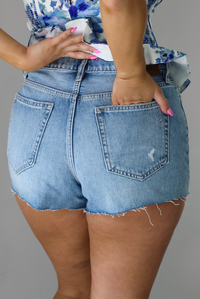 Woman wearing a light-wash pair of denim shorts that have distressed detailing. The photo shows the back of the shorts. The photo is taken indoors against a white wall.