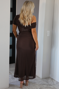 Woman model wearing a chocolate brown formal style dress that's fitted at the bodice, has asymmetrical sleeves, and a mermaid style skirt that's fitted at the hips and flared from the bottom of the high to the hem. The photo is taken inside a modern home in an archway. The photo showcases the back of the dress.