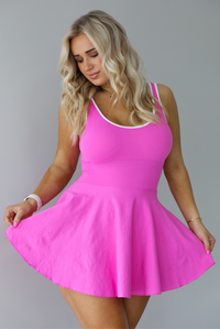Woman wearing a hot pink athleisure dress that has white trim, a fitted bodice, and flowy skirt. The photo shows a closeup of the dress. The photo is taken indoors against a white wall.