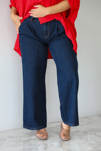 A woman wearing an oversized red top and dark wash denim jeans while standing indoors against a white wall with white marble flooring. 