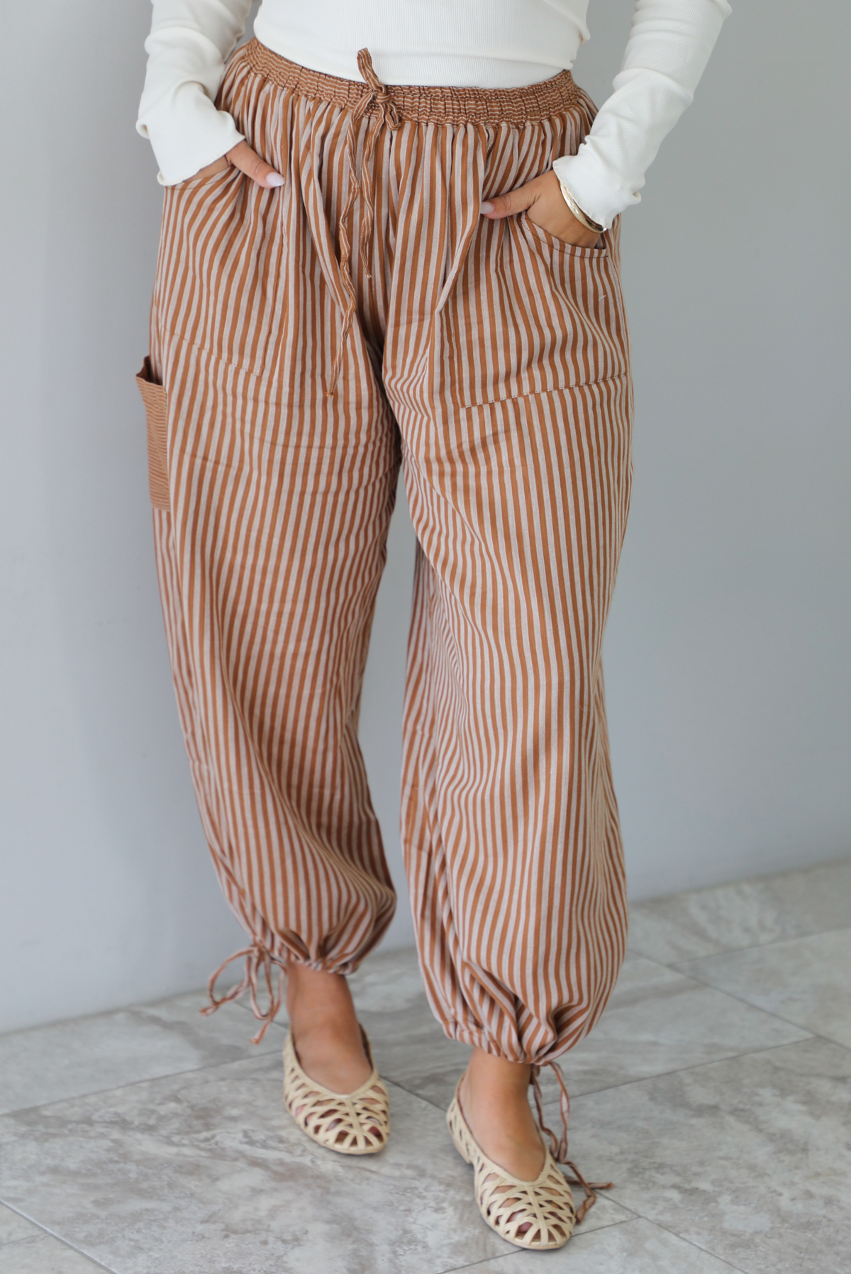 A closeup of a woman wearing a pair of camel and cream striped balloon pants that have an elastic and drawstring waistline with drawstring details at the bottom hem at the ankles, standing indoors against a white wall with white marble flooring. 