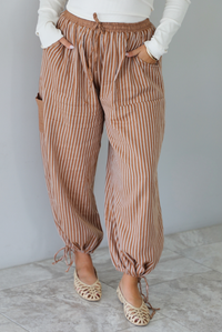 A closeup of a woman wearing a pair of camel and cream striped balloon pants that have an elastic and drawstring waistline with drawstring details at the bottom hem at the ankles, standing indoors against a white wall with white marble flooring. 