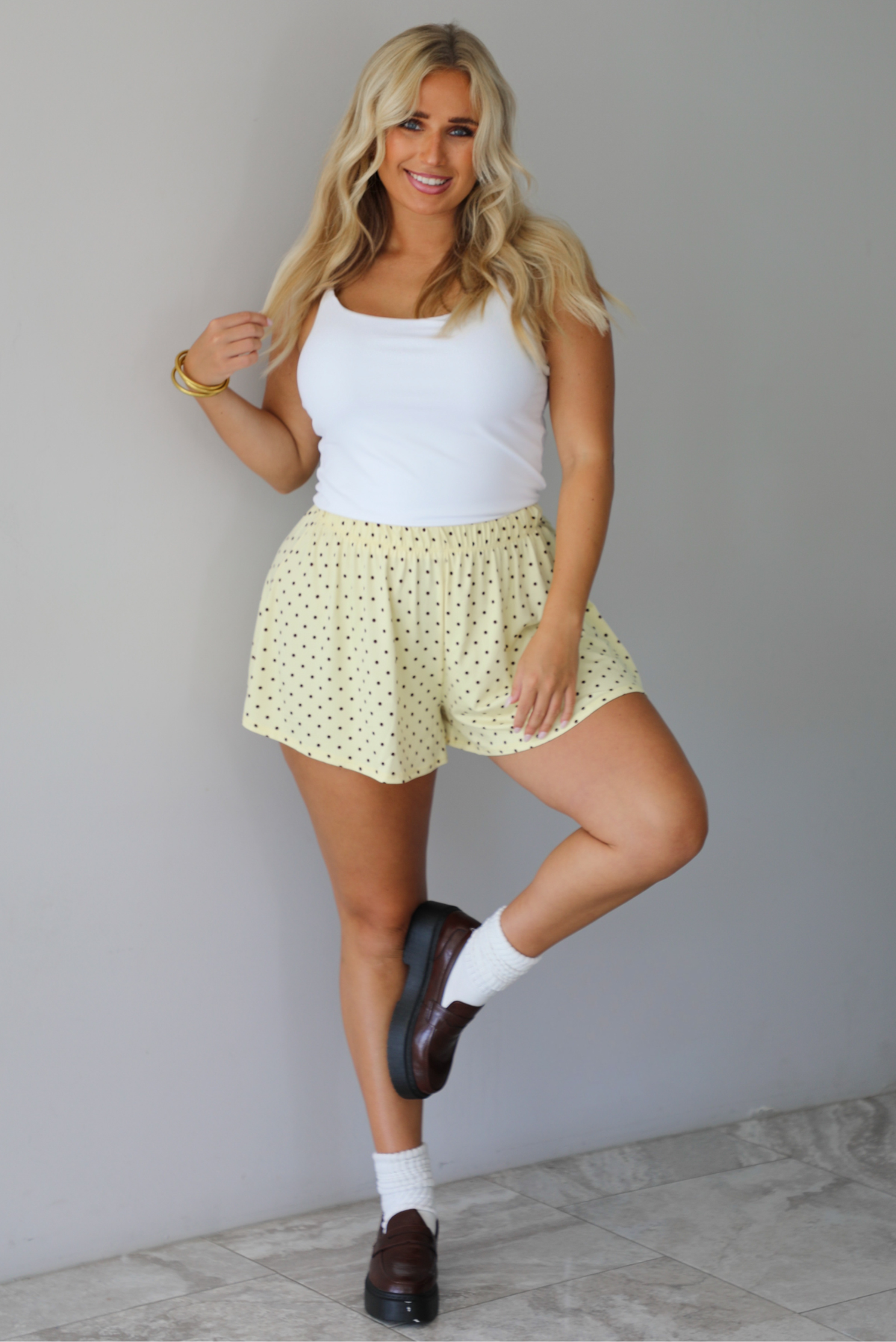 A woman modeling a white tank and pale yellow shorts that have a brown polka dot pattern throughout, standing indoors against a white wall with white marble flooring. 