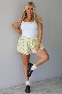 A woman modeling a white tank and pale yellow shorts that have a brown polka dot pattern throughout, standing indoors against a white wall with white marble flooring. 