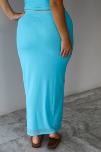 A blonde woman modeling a bright turquoise blue set with large pink and white hibiscus flower prints, indoors against a white wall with white marble flooring. The photo shows a closeup of the back of the skirt, which is high waisted with a maxi length, and does not have the floral pattern on it. 