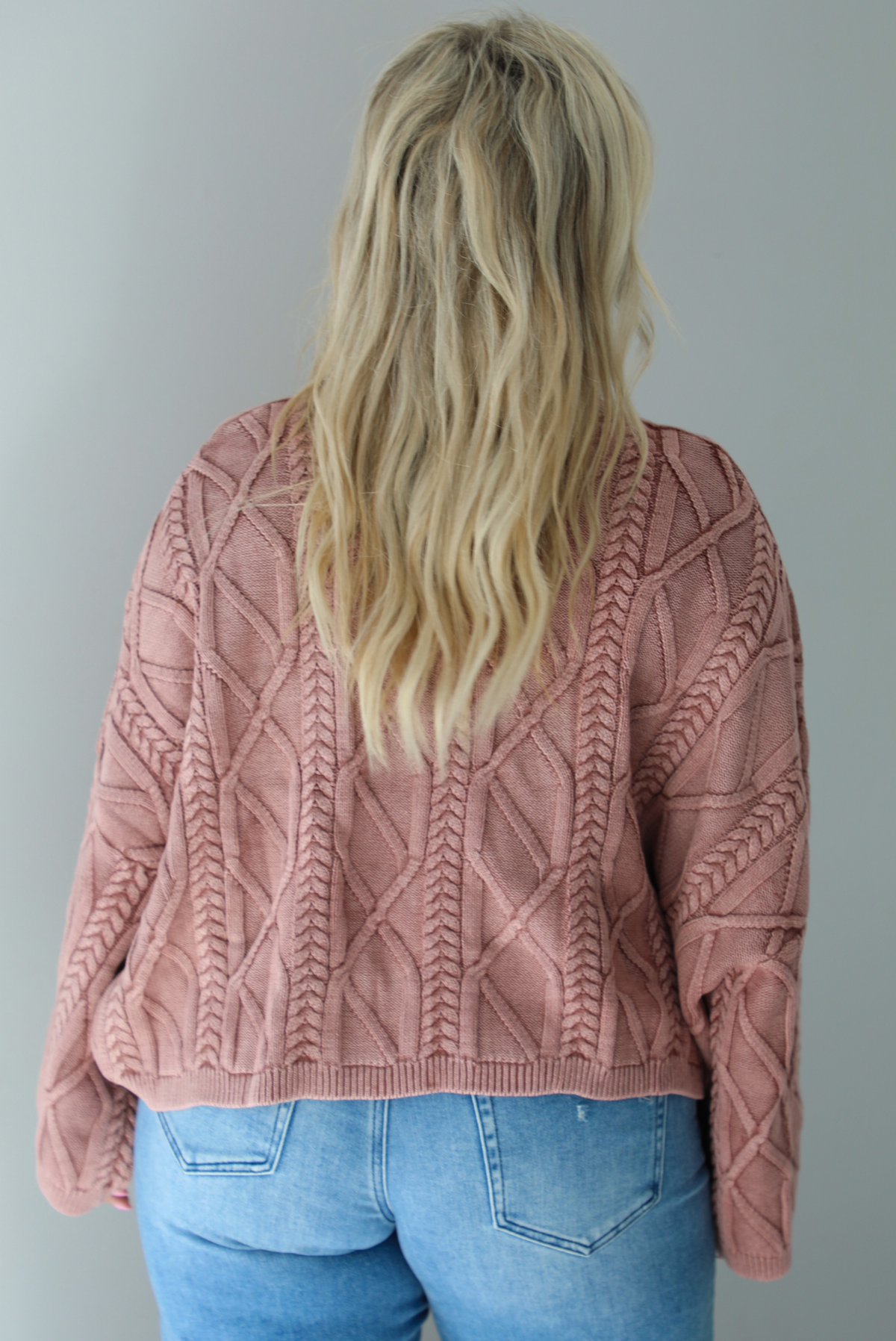 A woman shown from behind wearing a mulberry oversized sweater paired with a pair of medium wash distressed denim, standing indoors against a white wall.