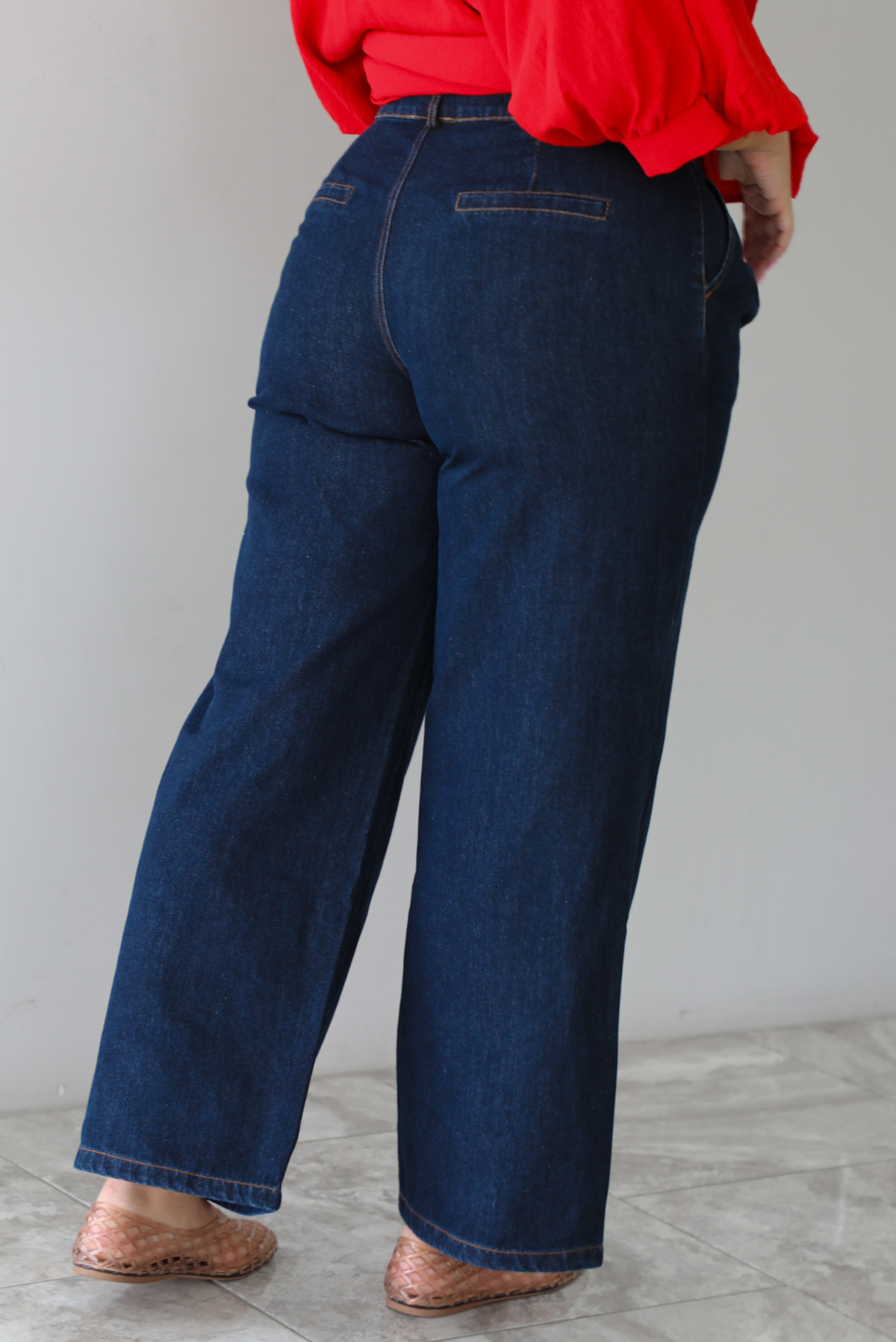 A woman shown from behind wearing an oversized red top and dark wash denim jeans while standing indoors against a white wall with white marble flooring. 