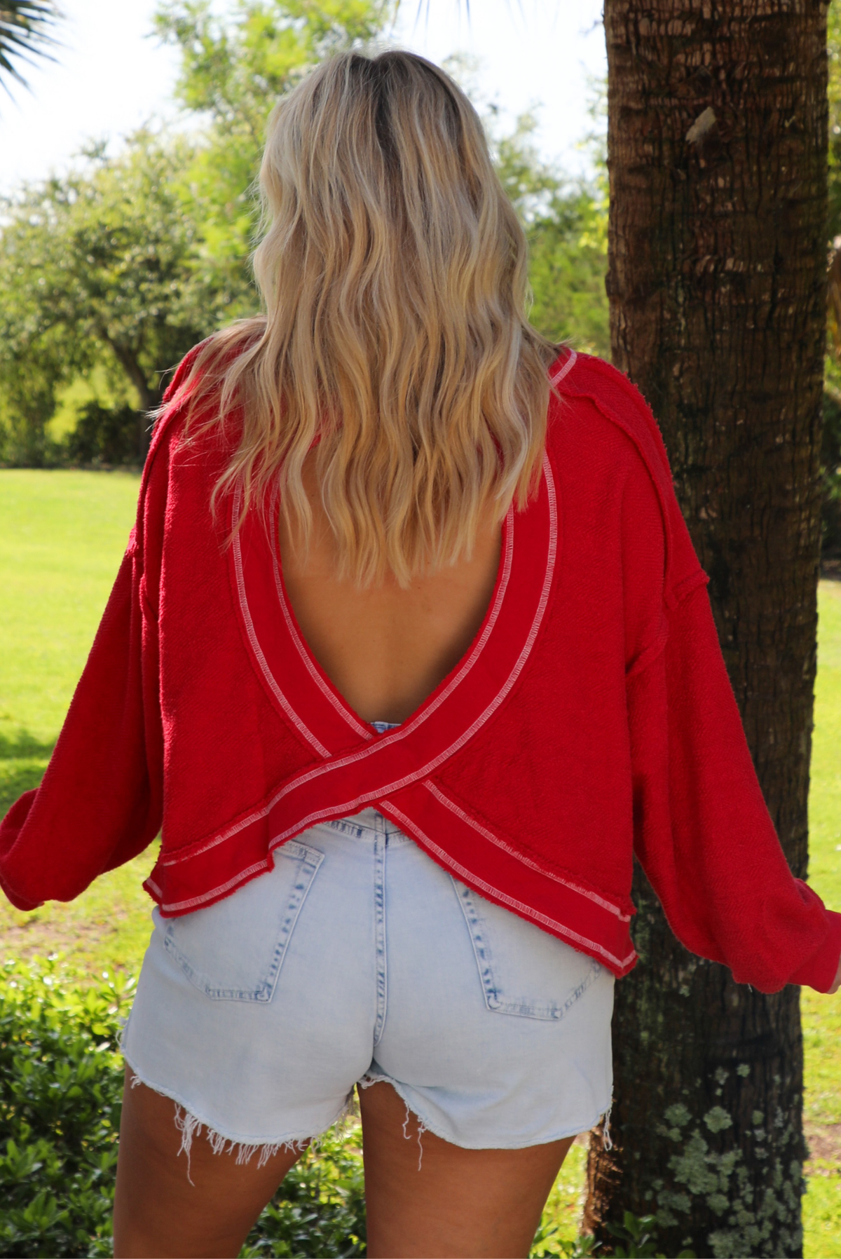 Woman wearing an oversized long sleeved sweater with light wash denim shorts outdoors with greenery in the background.  The photo shows the back of the sweater, which has an open back design.