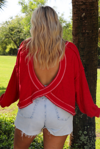 Woman wearing an oversized long sleeved sweater with light wash denim shorts outdoors with greenery in the background.  The photo shows the back of the sweater, which has an open back design.