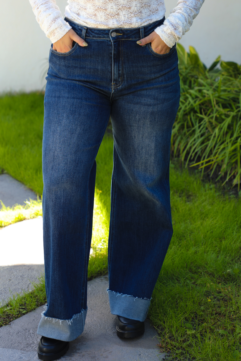 Woman model wearing dark denim blue jeans outdoors. They have a high waistline, functional button and zipper closures in the front, functional front and back pockets, and a slightly flared cut with a cuffed hem. 