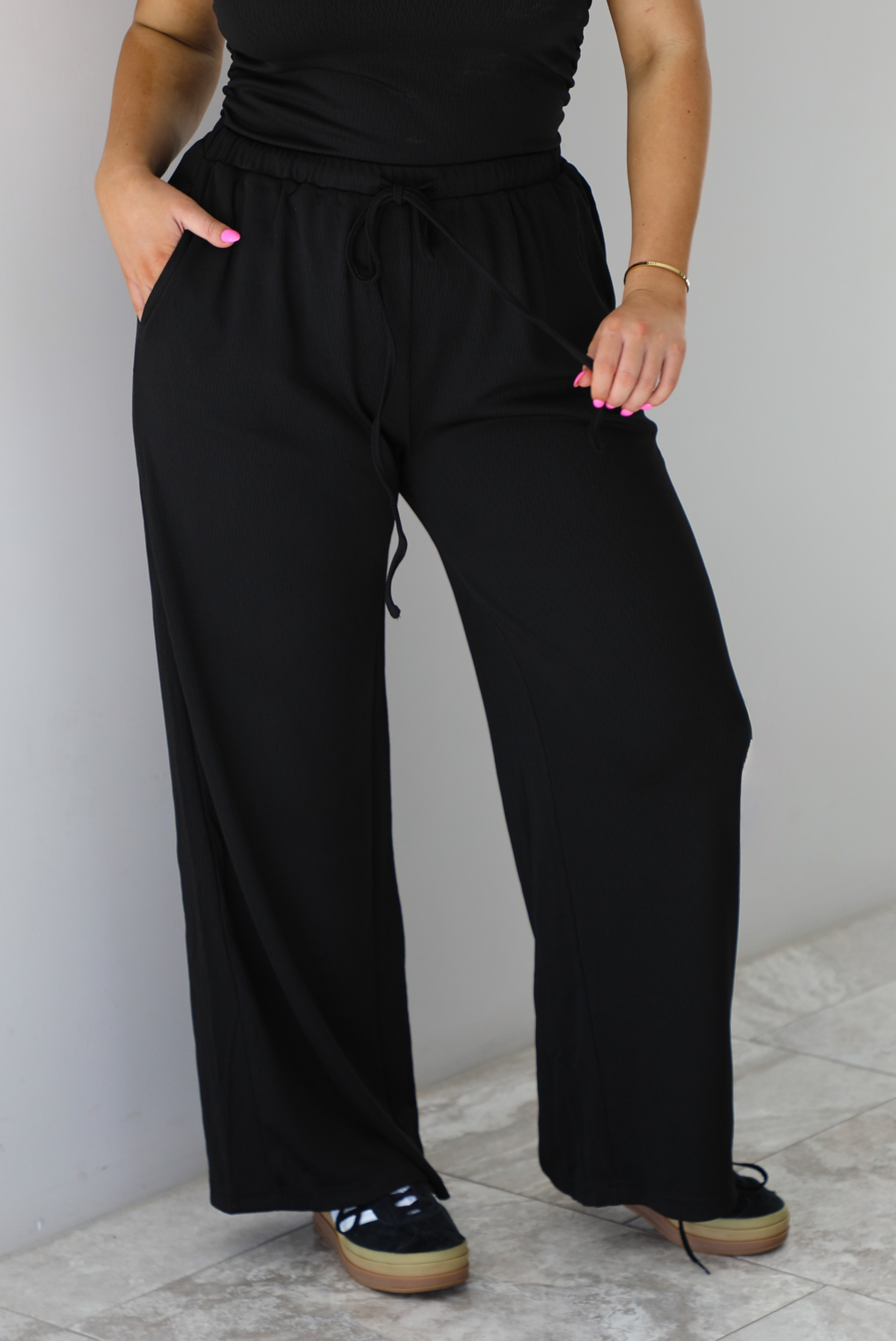 Woman wearing a two piece set featuring an off the shoulder top and high waisted pants. The photo shows a closeup of the pants.  The photo is taken indoors against a white wall with white marble flooring.