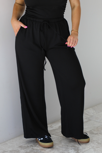Woman wearing a two piece set featuring an off the shoulder top and high waisted pants. The photo shows a closeup of the pants.  The photo is taken indoors against a white wall with white marble flooring.
